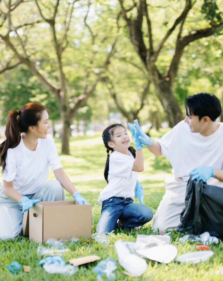A family wearing gloves is picking up trash in a sunny park. A child high-fives a parent, conveying teamwork and joy, with trees in the background.